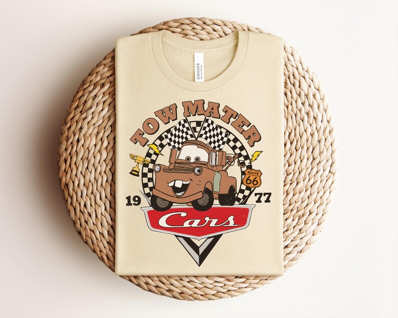 Puede incluir: Una camiseta de color crema con un gr&aacute;fico del personaje Tow Mater de la pel&iacute;cula Cars. El dise&ntilde;o incluye el texto Tow Mater, 1977, Route 66 y Cars, junto con banderas a cuadros, un trofeo y detalles de rayos.