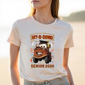 Op de afbeelding: Beige T-shirt met een cartoonafbeelding van Takel uit de film Cars, met een afstudeerhoed. De tekst "GIT-R-DONE!" staat boven Takel en "SENIOR 2026" staat eronder. Confetti en een geblokte vlag zijn ook inbegrepen.