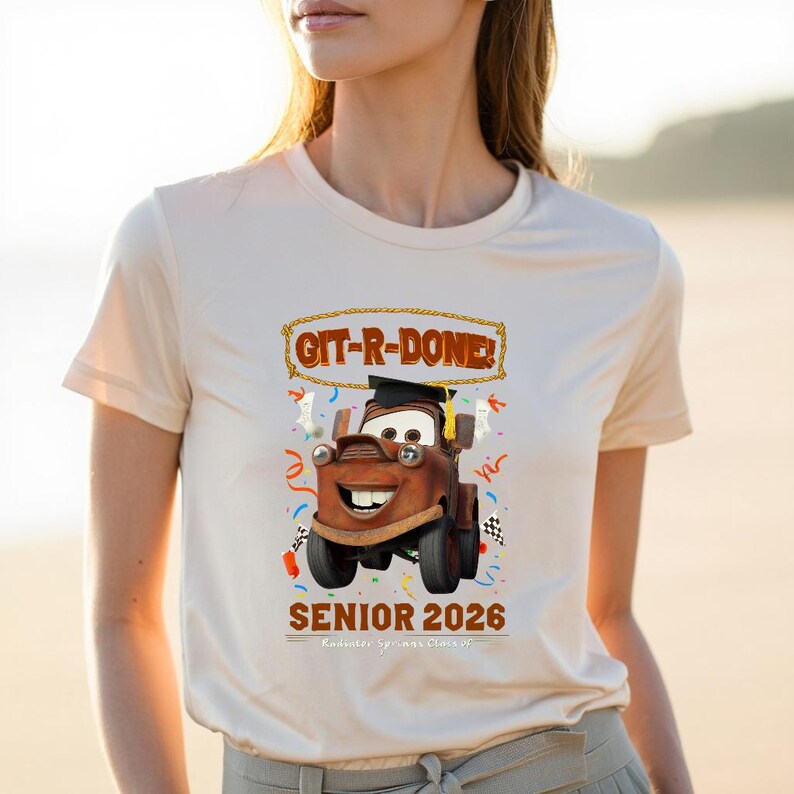 Op de afbeelding: Beige T-shirt met een cartoonafbeelding van Takel uit de film Cars, met een afstudeerhoed. De tekst "GIT-R-DONE!" staat boven Takel en "SENIOR 2026" staat eronder. Confetti en een geblokte vlag zijn ook inbegrepen.