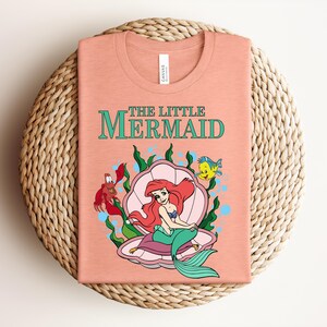 May include: A peach-colored t-shirt featuring a graphic of Ariel from The Little Mermaid sitting in a pink seashell. The design includes Sebastian the crab, Flounder the fish, green seaweed, and blue bubbles, with the text The Little Mermaid above.