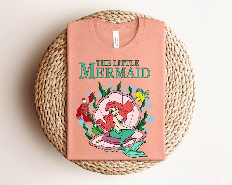 May include: A peach-colored t-shirt featuring a graphic of Ariel from The Little Mermaid sitting in a pink seashell. The design includes Sebastian the crab, Flounder the fish, green seaweed, and blue bubbles, with the text The Little Mermaid above.