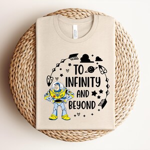 Peut inclure: T-shirt beige avec un dessin de Buzz l'&Eacute;clair et l'inscription noire "TO INFINITY and BEYOND". Le motif comprend des &eacute;toiles, des plan&egrave;tes et des &eacute;l&eacute;ments spatiaux. Le t-shirt est pli&eacute; sur une surface circulaire tiss&eacute;e.