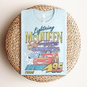 Puede incluir: Camiseta azul claro con Lightning McQueen y otros personajes de Cars. El gr&aacute;fico incluye el texto "Lightning McQueen" y "Think Fast 95". La camiseta est&aacute; doblada sobre una superficie circular tejida.