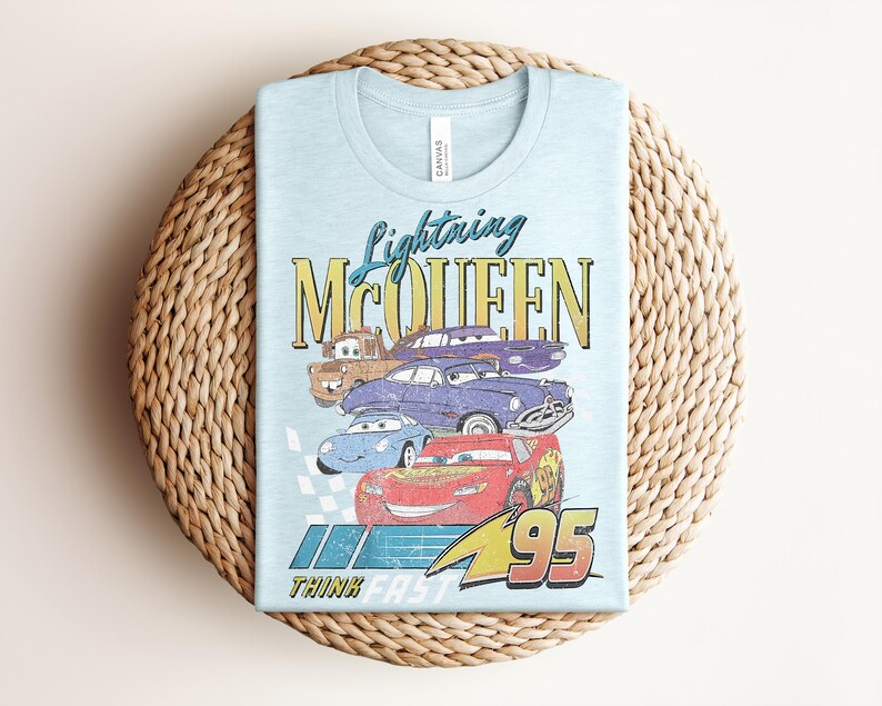 Puede incluir: Camiseta azul claro con Lightning McQueen y otros personajes de Cars. El gr&aacute;fico incluye el texto "Lightning McQueen" y "Think Fast 95". La camiseta est&aacute; doblada sobre una superficie circular tejida.