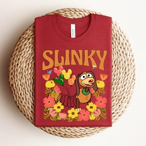 K&ouml;nnte beinhalten: Ein rotes T-Shirt mit dem Wort "SLINKY" in orangefarbenen Blockbuchstaben. Das Shirt hat eine Cartoon-Grafik eines Hundes mit Luftballons, Blumen und einer Biene. Das T-Shirt wird auf einem gewebten, runden Platzdeckchen pr&auml;sentiert.