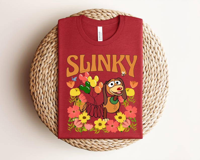 K&ouml;nnte beinhalten: Ein rotes T-Shirt mit dem Wort "SLINKY" in orangefarbenen Blockbuchstaben. Das Shirt hat eine Cartoon-Grafik eines Hundes mit Luftballons, Blumen und einer Biene. Das T-Shirt wird auf einem gewebten, runden Platzdeckchen pr&auml;sentiert.
