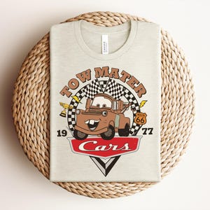 Puede incluir: Una camiseta de color beige claro con un estampado gr&aacute;fico del personaje Tow Mater de la pel&iacute;cula Cars. El dise&ntilde;o incluye una bandera a cuadros, un trofeo dorado, una se&ntilde;al de la Ruta 66, el texto Tow Mater, 1977 y el logo rojo Cars.