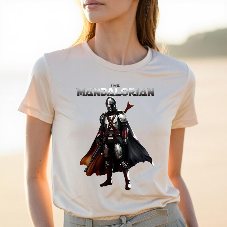 Op de afbeelding: Cr&egrave;mekleurig T-shirt met een afbeelding van het personage The Mandalorian. De afbeelding toont het personage in volledige uitrusting, met een zwarte cape en een rood wapen. De tekst "THE MANDALORIAN" staat boven het personage.