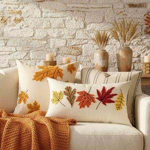 May include: A cream-colored square throw pillow with embroidered orange maple leaves and a rectangular lumbar pillow with embroidered multi-colored autumn leaves, both resting on a white sofa with a chunky knit orange throw blanket draped over it.