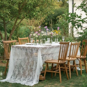 May include: An outdoor dining scene featuring a wooden table draped with a white lace tablecloth. Wooden chairs surround the table, which is adorned with small vases of wildflowers. The setting is a verdant garden.