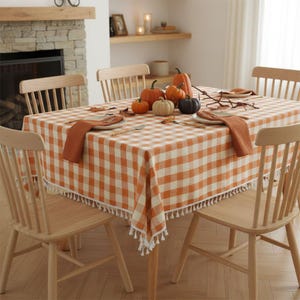 May include: A rectangular dining table covered with an orange and white chequered tablecloth featuring white fringe trim. The table is set with two orange cloth napkins, two white plates, and a centrepiece of five assorted pumpkins.
