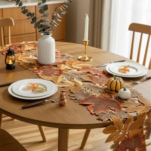 May include: An oval wooden dining table set for a fall-themed meal. A leaf-shaped table runner in shades of brown and orange runs down the center. White plates, a small pumpkin, and a candle holder are also on the table.