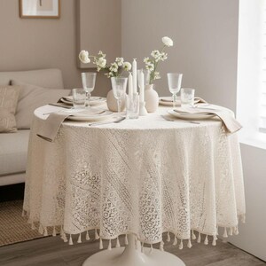 Könnte beinhalten: Ein runder Esstisch, der für eine Mahlzeit gedeckt ist. Der Tisch ist mit einer cremefarbenen Spitzentischdecke mit Quastenbesatz bedeckt. Der Tisch ist mit Tellern, Gläsern, Besteck, Servietten, Kerzen und Blumen gedeckt.