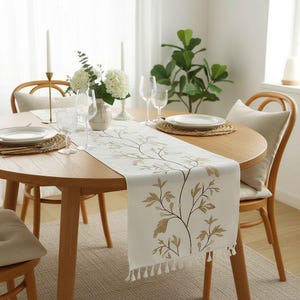 Könnte beinhalten: Cremefarbener Tischläufer mit einem botanischen Design aus braunen Blättern und Zweigen. Der Läufer liegt auf einem runden Holztisch, der für eine Mahlzeit gedeckt ist, mit Tellern, Gläsern und Kerzen. Holzstühle.