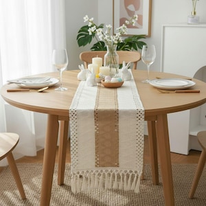 Peut inclure: Une table à manger ronde en bois décorée pour Pâques. Un chemin de table crème et marron avec des franges est centré sur la table. Des figurines de lapin en céramique blanche, des bougies et un bol d'œufs de Pâques sont disposés sur le chemin de table.