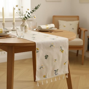 Può includere: Runner da tavolo in lino beige con ricami floreali gialli e verdi. Il runner ha una frangia di nappe bianche all'estremità. Il runner è posto su un tavolo di legno, con un vaso di fiori e candele sullo sfondo.