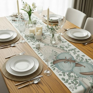 Puede incluir: Una mesa de comedor de madera preparada para una ocasión especial. La mesa está decorada con un camino de mesa floral, platos blancos, cubiertos, velas y copas de vino. El camino de mesa presenta huevos de Pascua azules.