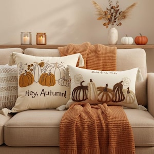 Könnte beinhalten: Zwei cremefarbene Dekokissen mit strukturierter Kürbisstickerei und Quasten an den Ecken. Ein quadratisches Kissen trägt den Text "Hey, Autumn!" und das rechteckige Kissen zeigt das Wort "autumn" in kursiver Schrift.