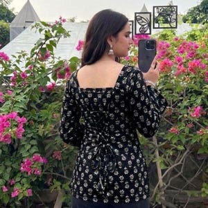 Può includere: Top nero con stampa floreale bianca, scollatura quadrata e maniche lunghe. La schiena ha un design a corsetto con lacci. La persona tiene in mano un telefono. Il capo fa parte di una collezione di abbigliamento.