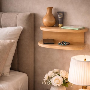 Può includere: Un comodino sospeso in legno chiaro a due ripiani. Un vaso di legno, un tubetto di lozione e un piccolo libro sono sul ripiano superiore. Uno smartphone e un braccialetto di perline sono sul ripiano inferiore. Un bouquet di rose bianche è sotto.