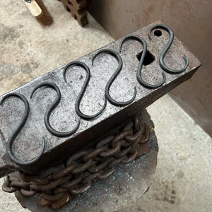 Puede incluir: Cinco ganchos en S de metal oscuro hechos a mano descansan sobre un yunque desgastado. Los ganchos están espaciados uniformemente y tienen un diseño simple y curvo. Una cadena pesada sostiene el yunque, lo que sugiere un tema de herrería.