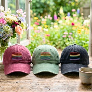May include: Three baseball caps in burgundy, olive green, and black, each with a rainbow-colored embroidered patch. The caps are displayed on a wooden surface, with a window and flowers in the background.