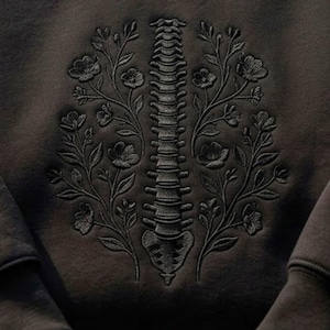 Op de afbeelding: Een zwart sweatshirt met een ton-sur-ton zwarte geborduurde afbeelding van een menselijke ruggengraat, verticaal gecentreerd. De ruggengraat wordt aan beide zijden geflankeerd door symmetrische, gedetailleerde bloementakken met bladeren en bloemen.