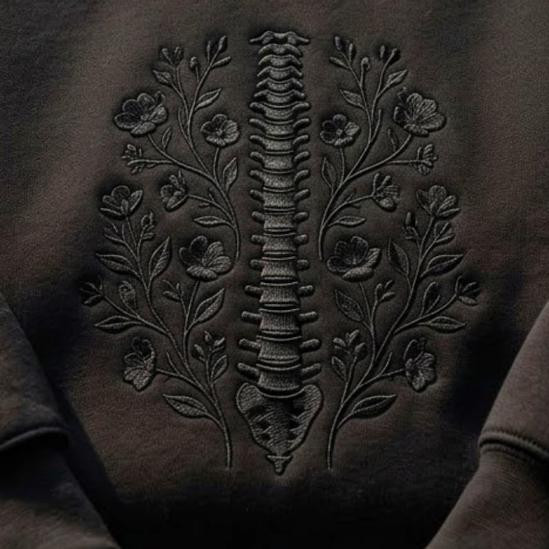 Op de afbeelding: Een zwart sweatshirt met een ton-sur-ton zwarte geborduurde afbeelding van een menselijke ruggengraat, verticaal gecentreerd. De ruggengraat wordt aan beide zijden geflankeerd door symmetrische, gedetailleerde bloementakken met bladeren en bloemen.