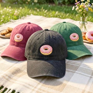 Puede incluir: Tres gorras de béisbol en color granate, negro y verde, cada una con un dónut rosa bordado con chispas de colores en el panel frontal. Las gorras tienen una corona estructurada, una visera curva y una textura de tela desgastada.