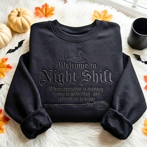May include: A black crewneck sweatshirt featuring embroidered text that reads Welcome to Night Shift, Where everyday is monday, today is yesterday, and torromow is today, accompanied by a witch hat icon and a broomstick graphic.
