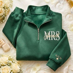 May include: A dark green quarter-zip sweatshirt featuring a silver metal zipper. The chest displays the white text "MRS" with the name "Jarrell" in gold script. The left sleeve cuff is embroidered with the date "11.15.26" and a small gold heart.