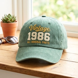 Könnte beinhalten: Eine salbeigrüne Baseballkappe mit verwaschener Optik und der gestickten Aufschrift Vintage 1986 ALL ORIGINAL PARTS. Die Kappe verfügt über einen gebogenen Schirm, ein strukturiertes Vorderteil und gestickte Ösen zur Belüftung.
