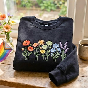 May include: A black crewneck sweatshirt featuring a colorful embroidered design of various wildflowers across the chest. The embroidery includes orange poppies, yellow daisies, light green blossoms, blue flowers, and purple lavender sprigs with green stems.