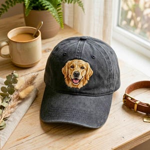 Op de afbeelding: Een zwarte baseball cap met een geborduurd beeld van een golden retriever. De hond heeft een blije uitdrukking. De pet ligt op een houten oppervlak, met een koffiemok en een hondenriem in de buurt.