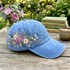 May include: A light blue baseball cap with embroidered floral design. The embroidery features pink, purple, and cream-coloured flowers with green leaves. The cap is sitting on a wooden surface.