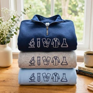 May include: A stack of three quarter-zip sweatshirts in navy blue, heather grey, and light blue. Each sweatshirt features a white or dark blue line art graphic of a microscope, test tube, two slides, a lab coat, and an Erlenmeyer flask.