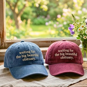 May include: Two baseball caps, one blue and one burgundy, are displayed. Each cap has the phrase "waiting for the big beautiful obituary" embroidered in white. The caps are on a neutral-colored surface, with a window and flowers in the background.