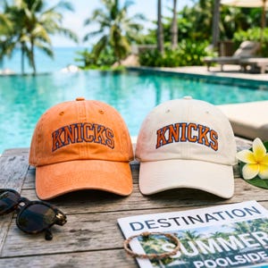 Puede incluir: Dos gorras de béisbol, una naranja y otra color crema, cada una con la palabra KNICKS bordada en letras arqueadas azules y naranjas en la parte frontal. Las gorras tienen una corona estructurada, una visera curva y un botón en la parte superior.