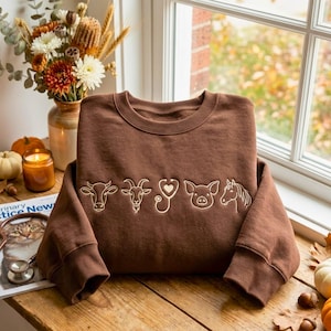 May include: A brown crewneck sweatshirt featuring a white line art graphic across the chest. The design includes a cow, a goat, a stethoscope with a heart-shaped chest piece, a pig, and a horse arranged in a horizontal row on the fabric.