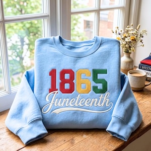 Op de afbeelding: Een lichtblauw sweatshirt met het jaartal "1865" in rood, geel en groen, en het woord "Juneteenth" in witte letters. Het sweatshirt ligt op een houten oppervlak bij een raam.