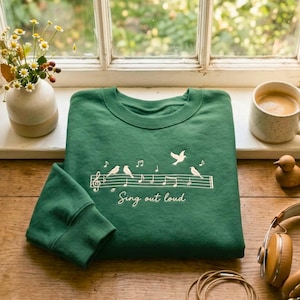 May include: A forest green crewneck sweatshirt featuring white embroidery of a musical staff with birds perched on the lines, flying birds, musical notes, and the text "Sing out loud" written in a cursive script across the centre chest.