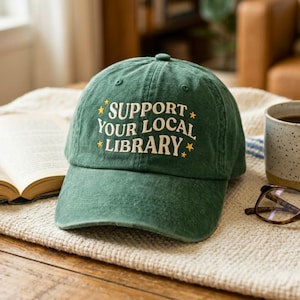 May include: A green baseball cap with the text "SUPPORT YOUR LOCAL LIBRARY" in white and yellow. The hat is sitting on a table next to a book, a cup of coffee, and glasses.