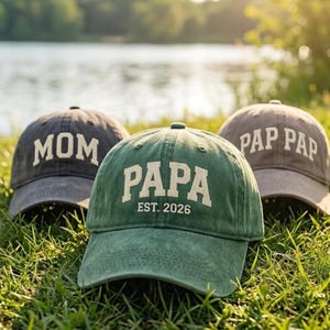 Puede incluir: Tres gorras de béisbol en verde, gris oscuro y marrón claro. La gorra verde presenta el texto "PAPA EST. 2026" en blanco. La gorra gris oscuro muestra la palabra "MOM" en blanco y la gorra marrón claro tiene el texto "PAP PAP" en blanco.