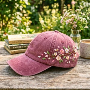 May include: A maroon baseball cap with embroidered floral detailing. The cap features a curved brim and a six-panel design. The floral embroidery includes pink, white, and green flowers and leaves, adding a touch of nature-inspired style.