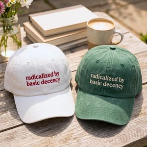 Puede incluir: Dos gorras de béisbol, una blanca y otra verde, se exhiben sobre una superficie de madera. Ambas gorras tienen la frase "radicalized by basic decency" bordada en la parte delantera. Una taza de café, libros y flores están en el fondo.