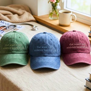 May include: Three baseball caps in green, blue, and maroon, each featuring a distressed fabric finish and the embroidered text "that wasn't very evidence based of you" centered on the front panel above the curved brim of each hat.