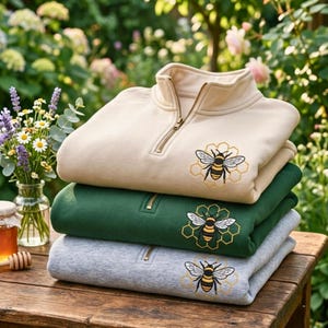 May include: A stack of three quarter-zip sweatshirts in beige, dark green, and heather gray. Each sweatshirt features an embroidered design of a honeybee centered over a yellow honeycomb pattern on the chest and a metal zipper at the collar.