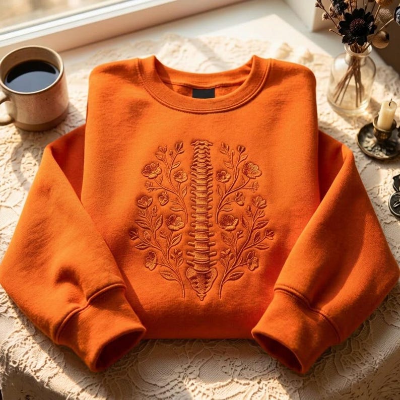 Op de afbeelding: Een feloranje sweater met ronde hals en een gedetailleerd, tonaal borduurontwerp op de voorzijde. Het borduurwerk toont een menselijke ruggengraat in het midden tussen twee symmetrische bloementakken met bloemen en bladeren.