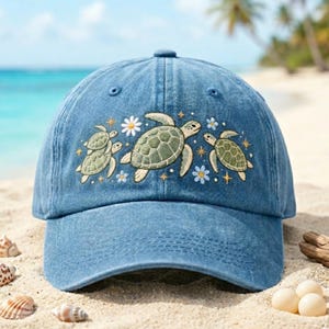 May include: A blue denim baseball cap featuring an embroidered design of three green sea turtles swimming among small white and blue flowers and gold star accents. The cap has a structured crown and a curved brim with a classic six-panel construction.