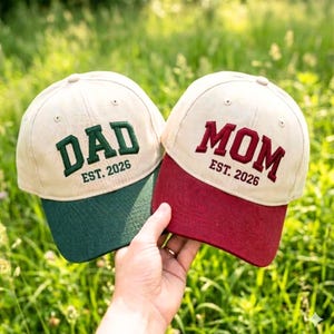 Peut inclure: Deux casquettes de baseball avec des couronnes beiges et des visières colorées sont tenues devant un fond vert. L'une a une visière verte et le mot "DAD" brodé en vert, avec "EST. 2026" en dessous. L'autre a une visière bordeaux et le mot "MOM" brodé en bordeaux, avec "EST. 2026" en dessous.